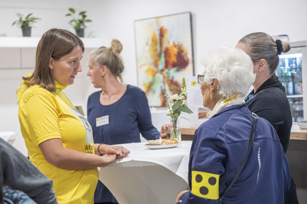 Frau mit gelbem Shirt redet mit älterer Frau mit gelber Armkennzeichnung