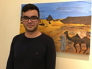 Praktikant Ariel steht vor einem Gemälde welches eine Wüste mit Kamelen zeigt