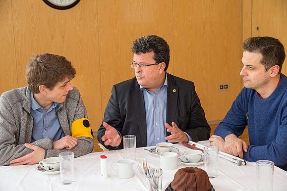 Drei Männer sitzen mit Kaffee und Kuchen bei Tisch und diskutieren