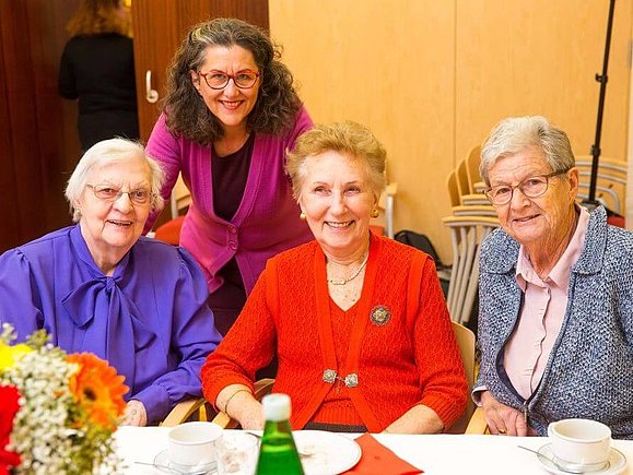 Frau J. sitzt bei einem Testatoren-Treffen mit zwei weiteren Damen an einem Tisch, im Hintergrund ist die Mitarbeiterin der Hilfsgemeinschaft, Frau Portner-Frisch zu sehen.