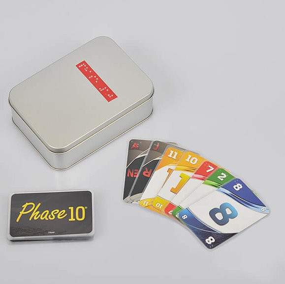 Eine Metallbox mit Braille-Schrift, eine Karte mit dem Phase 10 Logo und mehrere farbige Zahlenkarten mit Prägung liegen am Tisch.