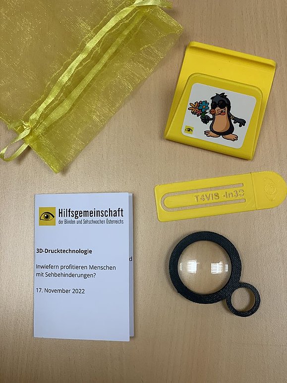 Gelbes Täschchen, schwarze kleine Lupe, gelber Handystehhalter mit Maulwurfgrafik, gelbes Lesezeichen und Papierkärtchen mit Logo der Hilfsgemeinschaft 