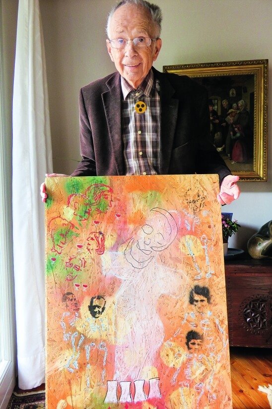 Ein Mann mit Brille und einem gelben Button mit 3 schwarzen Punkten steht in einem Wohnzimmer und hält ein buntes Gemälde vor sich.