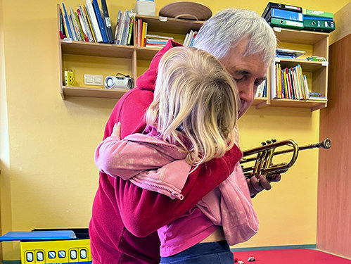 Älterer Mann und kleines Mädchen umarmen sich in einem Spielzimmer, er kniet und hält ein Blasinstrument in der Hand.