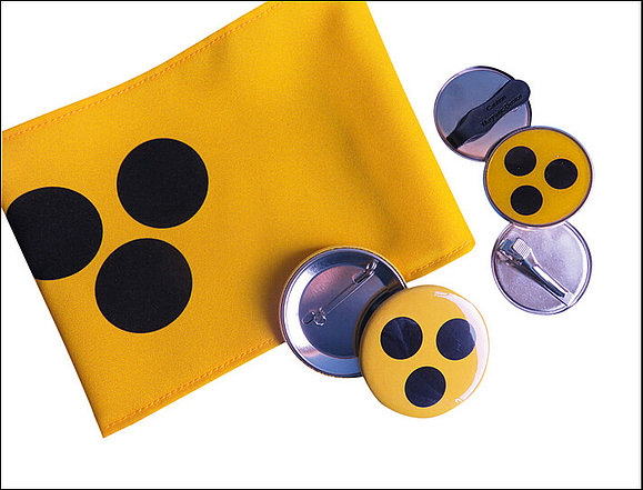 Gelbe Armschleife mit drei schwarzen Punkten und fünf kleine gelbe Plaketten mit drei schwarzen Punkten zum Anstecken mit Nadel oder Clip