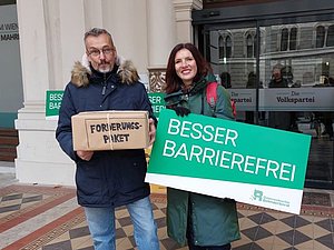 Mann mit Kiste "Forderungspaket" und Frau mit grünem Schild "Besser barrierefrei" vor Hauseingang