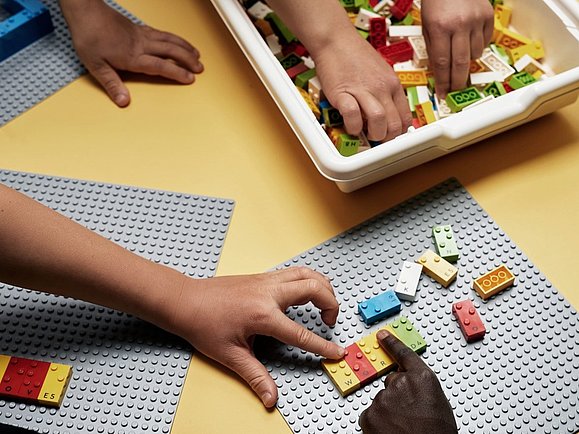 Drei Paar Kinderhände greifen zu bunten Lego Steinen, die auf einem grauen Steckbrett stecken. Die Noppen der Legosteine entsprechen Buchstaben der Braille Schrift. Die jeweiligen Buchstaben sind auch in Schwarz auf den Steinen aufgedruckt. 