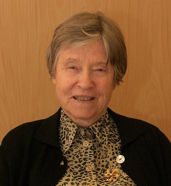 Ältere Frau mit Kurzhaarfrisur und Leoprinthemd und schwarzer Jacke.