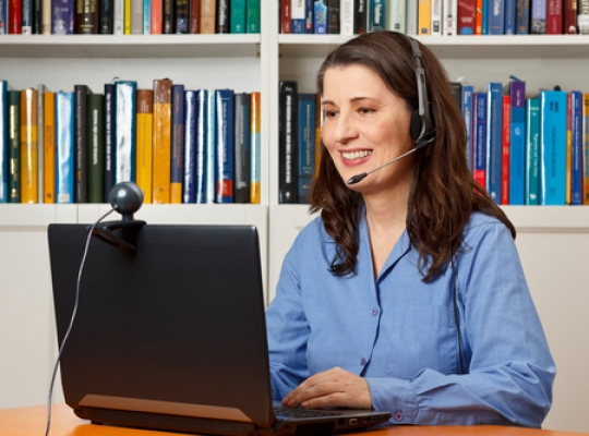 Beratung mittels Video-Telefonie Eine junge Frau sitzt mit einem Headset auf dem Kopf vor einem Laptop, dieser hat eine Kamera befestigt.