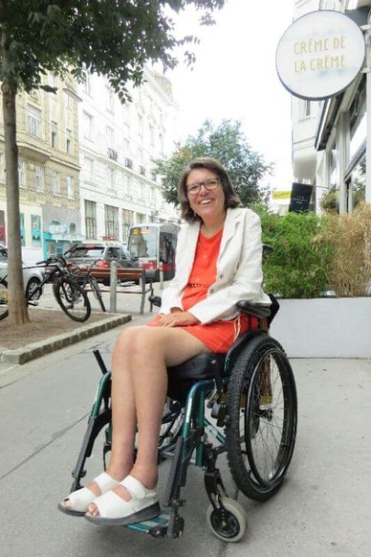 Expertin Maria Grundner trägt ein orangenes Kleid mit weißem Blazer und sitz in einem Rollstuhl.