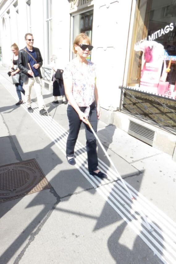 Frau mit Langstock gleitet am Blindenleitsystem entlang.