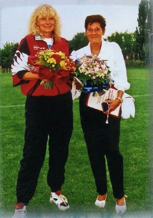 Frau in Sportkleidung und Frau mit weißer Bluse stehen mit Blumenstrauß in der Hand auf einem Fußballfeld