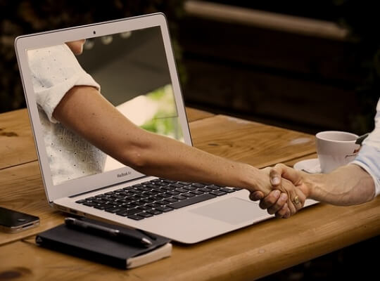 Laptop bei dem ein Arm aus dem Display hinauskommt und einer anderen Person die Hand Schüttelt