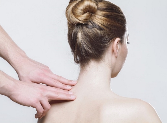Nackter Rücken einer Frau, zwei Hände deuten auf die Halswirbelsäule.