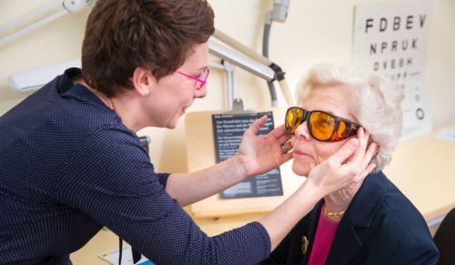 Mitarbeiterin der Hifsgemeinschaft setzt einer älteren Dame eine Kantenfilter-Brille auf