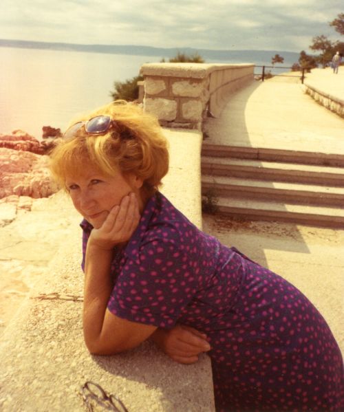 Auf einem leicht vergilbten Foto sieht man eine Frau in einer mediterranen Landschaft am Meer, an einer Mauer lehnend in die Kamera lächeln.