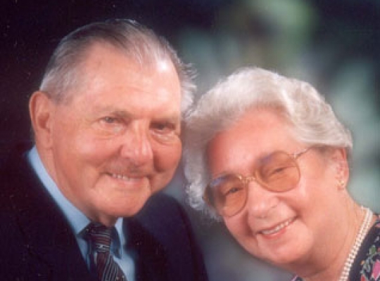 Ein älteres Ehepaar sitzt einander zugeneigt und lächelt fröhlich in die Kamera
