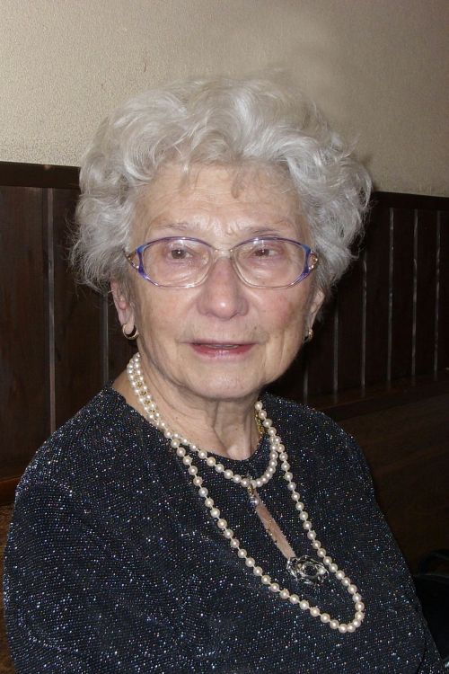 Eine ältere Dame mit Dauerwelle, großer Brille und mehrerer Ketten um den Hals blickt leicht lächelnd in die Kamera