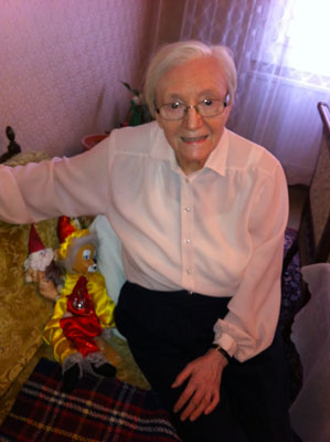 Eine ältere Dame sitzt fröhlich lächelnd auf einem Sofa, neben ihr bunte Puppen