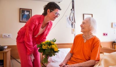 Mitarbeiterin Alexandra Kleinheinz bei einem Besuchsdienst im Krankenhaus, Copyright: Franz Pfluegl Eine Testamentsspenderin sitzt auf einem Bett. Von links beugt sich eine Mitarbeiterin mit einem Blumenstrauß zu ihr hin.