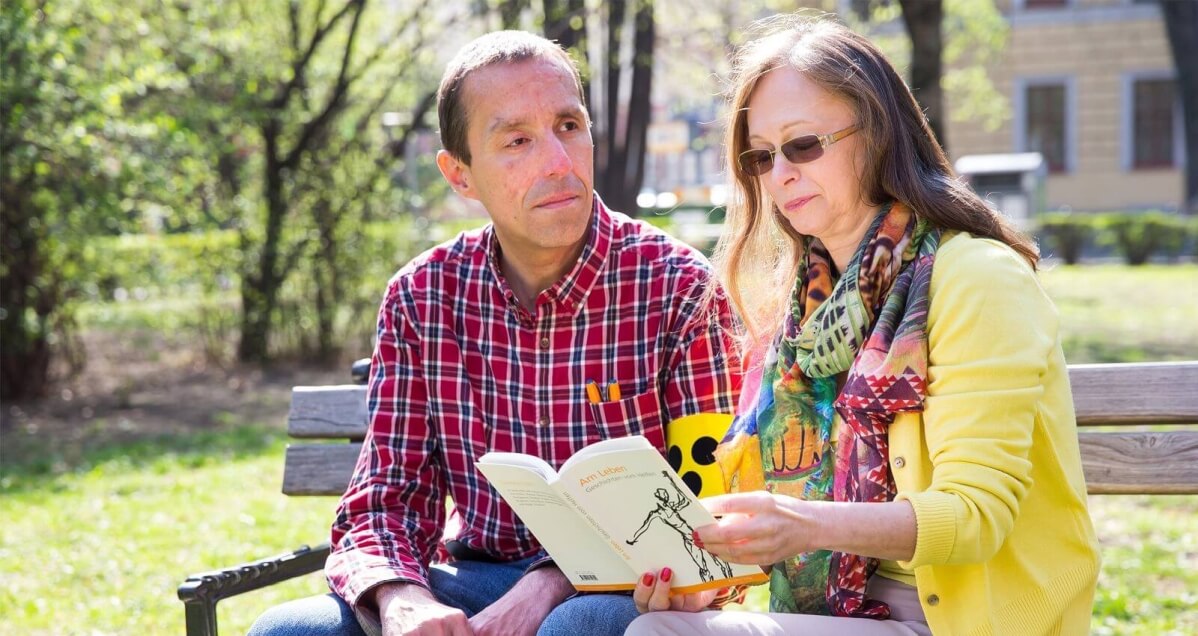 Eine Frau liest einem blinden Mann mit Armbinde ein Buch vor. Sie sitzen nebeneinander auf einer Parkbank.