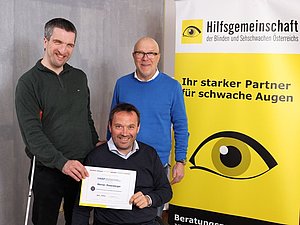 Drei Männer lächeln in die Kamera mit Zertifikat in der Hand vor gelbem Roll-Up mit Hilfsgemeinschaft Logo