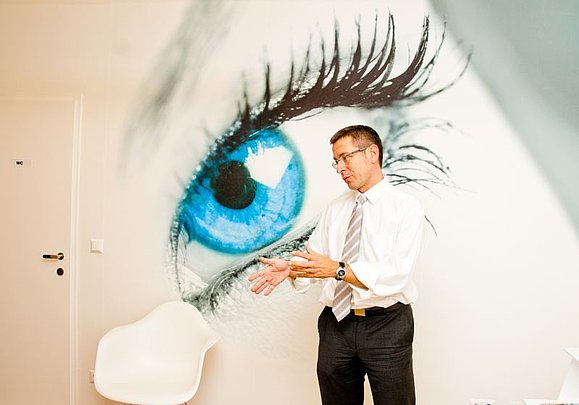 Ein Mann mit Brille steht gestikulierend vor einer Wand auf der ein großes blaues Auge zu sehen ist