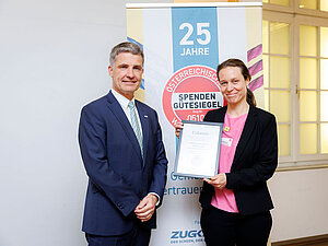 Foto: © Fundraising Verband Austria Eine Frau und ein Mann stehen vor einem Rollup worauf steht: "25 Jahre Spendengütesiegel". Die Frau hält eine Urkunde.