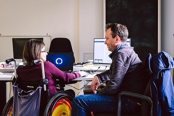 Eine junge Dame im Rollstuhl sitzt an ihrem Arbeitsplatz vorm PC. Neben ihr sitzt ihr persönlicher Assistent.