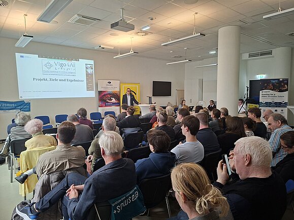Blick über die dicht besetzten Stuhlreihen eines Veranstaltungsraums auf einen Vortragenden. Neben ihm zeigt eine Wandprojektion die Präsentation „VIgo4AI – Projekt, Ziele und Ergebnisse“ zum Thema Menschen mit Sehbeeinträchtigungen und künstliche Intelligenz.