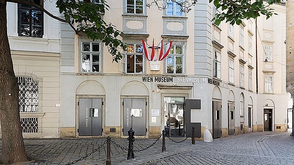 Ein mehrstöckiges Haus mit österreichischer Fahne und der Aufschrift "Wien Museum Uhrenmuseum". Davor ist ein kleiner Vorplatz mit einem Baum. 