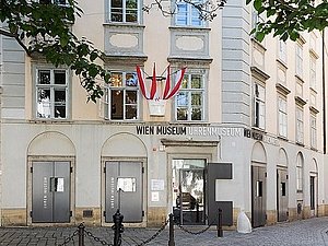 Ein mehrstöckiges Haus mit österreichischer Fahne und der Aufschrift "Wien Museum Uhrenmuseum". Davor ist ein kleiner Vorplatz mit einem Baum. 