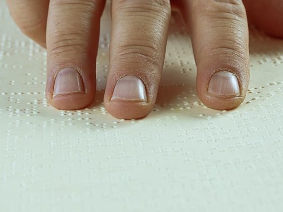 Drei Finger ertasten die Punkte der Brailleschrift auf einem Papier.