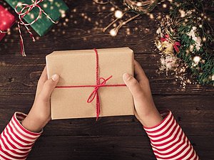 Hände halten braun eingepacktes Geschenk mit roter Masche über dunkelbraune Holzfläche