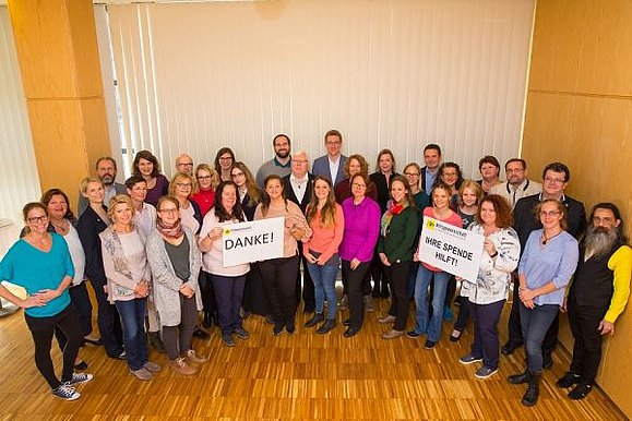 Die Mitarbeiter der Hilfsgemeinschaft stehen in einem Halbkreis in einem Raum nebeneinander. Zwei Schilder werden hoch gehalten. Eines mit "Danke" Aufschrift, ein weiteres mit "Ihre Spende hilft!". 