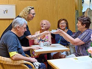 Ein Mann und drei Frauen sitzen bei einem Tisch. Eine weitere Frau steht und gibt der sitzenden Frau einen Kaffee. Am Tisch stehen Kaffeetassen und an einem Tisch im Vordergrund stehen rosa Blumen..