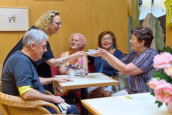 Ein Mann und drei Frauen sitzen bei einem Tisch. Eine weitere Frau steht und gibt der sitzenden Frau einen Kaffee. Am Tisch stehen Kaffeetassen und an einem Tisch im Vordergrund stehen rosa Blumen..