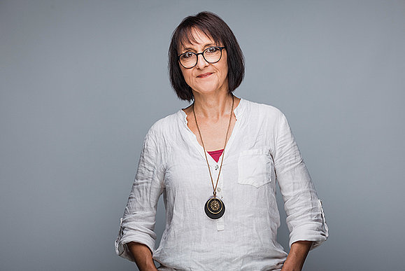 Standortleiterin Elke Zach-Tassati Eine Frau mit kurzen dunklen Haaren und Brille. Sie trägt eine weiße Bluse und eine Kette mit großem Anhänger und lächelt in die Kamera.
