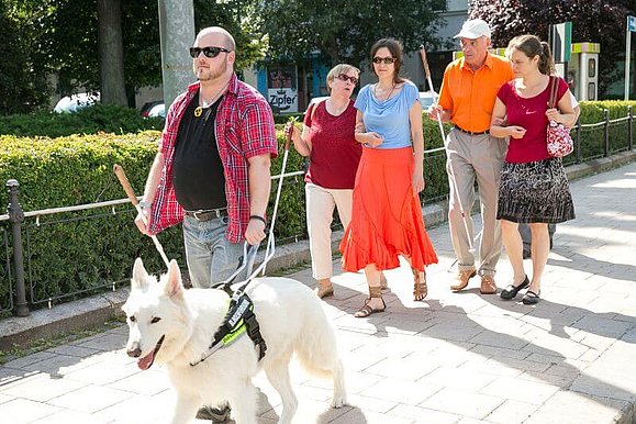 Ein Mann geht mit Blindenführhund voran, dahinter führen je zwei Menschen zwei blinde Menschen.