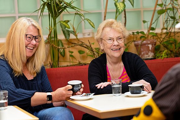 Zwei Frauen, eine mit langen blonden Haaren und eine älter mit kurzen blonden Haaren lachen und trinken gemeinsam Kaffee