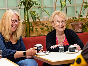 Zwei Frauen, eine mit langen blonden Haaren und eine älter mit kurzen blonden Haaren lachen und trinken gemeinsam Kaffee