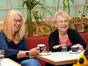 Zwei Frauen, eine mit langen blonden Haaren und eine älter mit kurzen blonden Haaren lachen und trinken gemeinsam Kaffee