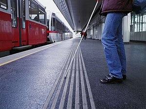 Person mit Langstock steht an Straßenbahnhaltestelle vor taktilem Bodenleitsystem und haltender Straßenbahn