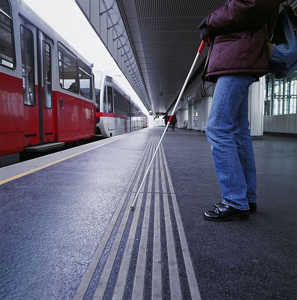 Person mit Langstock steht an Straßenbahnhaltestelle vor taktilem Bodenleitsystem und haltender Straßenbahn