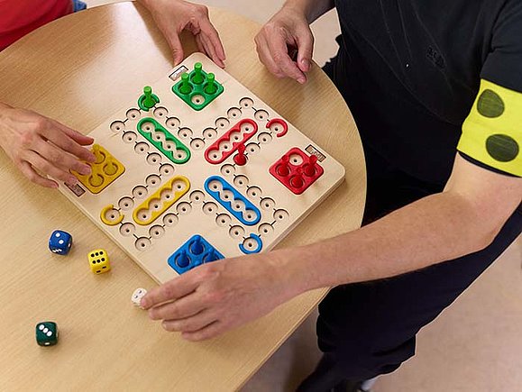 Blick von oben auf zwei Menschen, die Mensch-ärgere-dich-nicht auf einem taktilen Spielbrett spielen. Eine der Personen trägt eine gelbe Armbinde mit drei schwarzen Punkten. 