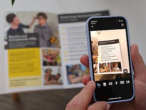 Smartphone im Fokus mit Vorleseapp, die gerade Infobroschüre mit der Kamera zoomt