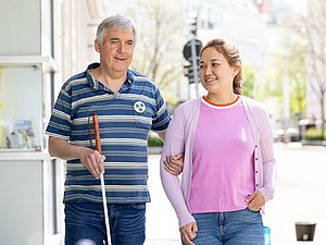 älterer Mann mit blaugestreiftem Shirt und weißem Stock wird von junger Frau mit lila Shirt geführt