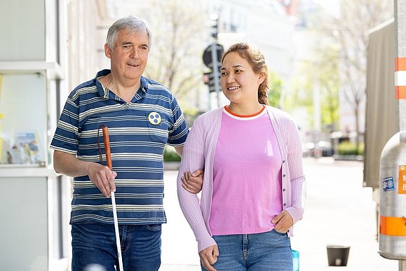 älterer Mann mit blaugestreiftem Shirt und weißem Stock wird von junger Frau mit lila Shirt geführt