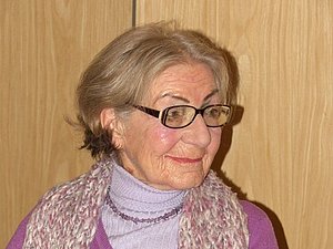 Eine ältere Dame mit Brille und Schal sitzt lächelnd an einem Tisch 