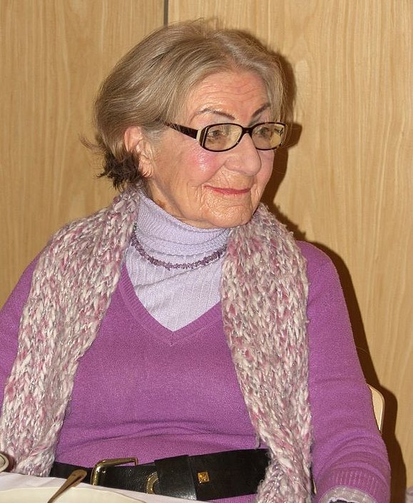 Eine ältere Dame mit Brille und Schal sitzt lächelnd an einem Tisch 
